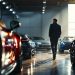 Un créateur automobile entouré de voitures de luxe dans un garage éclairé comme un showroom, ambiance éditoriale premium et lumineuse.