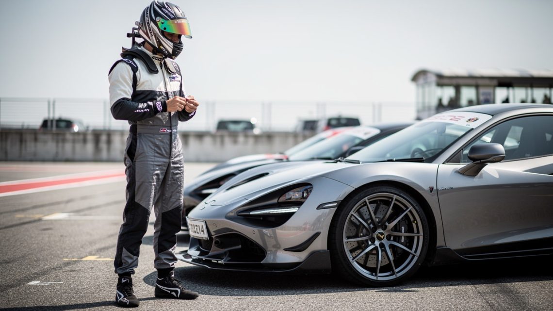 Un pilote amateur portant un casque intégral et une tenue couvrante se tient devant une voiture sportive de série dans le paddock d’un circuit, en pleine journée trackday.