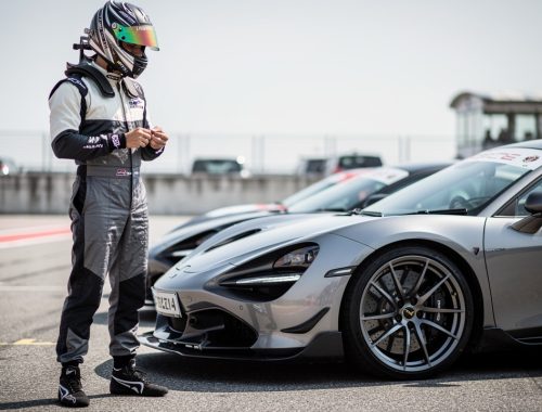 Un pilote amateur portant un casque intégral et une tenue couvrante se tient devant une voiture sportive de série dans le paddock d’un circuit, en pleine journée trackday.