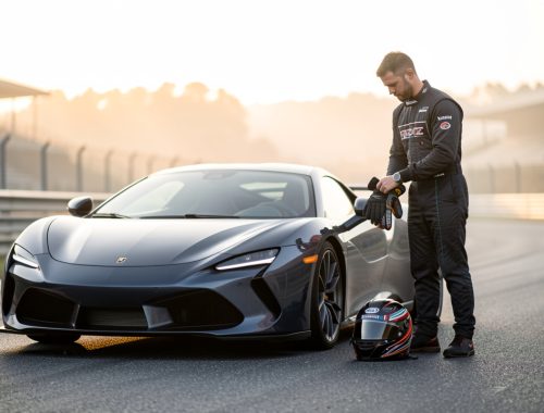 Une voiture sportive est garée dans un paddock de circuit tandis qu’un pilote prépare son casque et son équipement dans la lumière douce du matin.