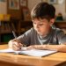 Un jeune garçon gaucher assis à un bureau d’école écrit au stylo dans un cahier sans spirale, dans une salle de classe lumineuse.