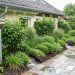 Jardin résidentiel lumineux après une pluie douce, avec récupérateur d’eau relié à une gouttière, noue végétalisée et allée en pavés drainants.